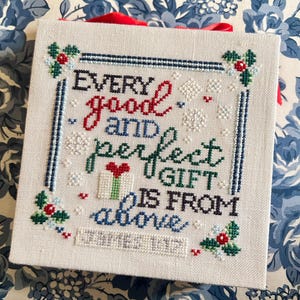 May include: Square, off-white cross-stitch with the text "Every good and perfect gift is from above." The text is in red, green, blue, and black. The border has holly and snowflake accents. A red ribbon is attached.