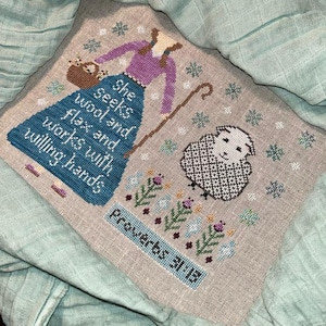 Puede incluir: Bordado de punto de cruz con una mujer con un suéter morado y un vestido azul que sostiene un cayado de pastor y una cesta de flores. El texto dice "Ella busca lana y lino y trabaja con manos dispuestas" y "Proverbios 31:13".