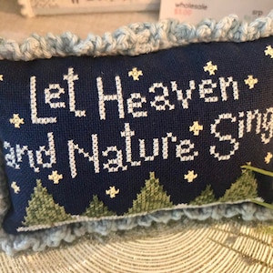 May include: A navy blue decorative pillow with the text "Let Heaven and Nature Sing" in white cross-stitch. The pillow features a light blue crocheted border and a design of green trees and yellow stars.