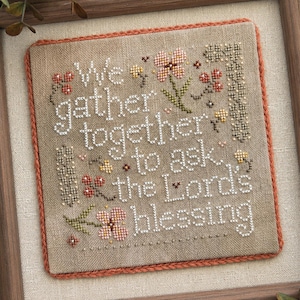 Pode incluir: Um mostruário de ponto cruz quadrado em tecido bege com o texto bordado em branco We gather together to ask the Lord's blessing. O design inclui pequenas flores rosas e amarelas, caules verdes e bagas vermelhas, emoldurado por uma borda cor de ferrugem.