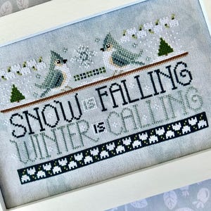 May include: Cross-stitch artwork in a white frame. The design features two blue birds perched on a branch above the words "SNOW IS FALLING WINTER IS CALLING." The artwork includes snowflakes, green trees, and a decorative border.