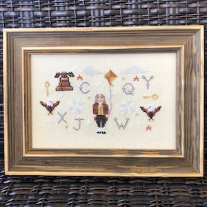 May include: A framed cross-stitch embroidery featuring a man holding a kite, a bell, and letters of the alphabet. The letters are X, J, W, C, Q, and Y. The embroidery is on a cream-colored fabric with brown and white accents.