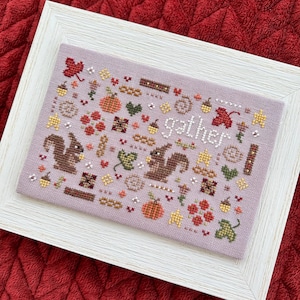 May include: A cross-stitch pattern with a light purple background featuring two brown squirrels, a pumpkin, and the word "gather". The pattern is surrounded by a white frame.