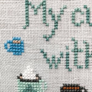 May include: Cross-stitch embroidery on white fabric with the words "my tea with" in green thread and images of teacups in blue, green, and gray thread.