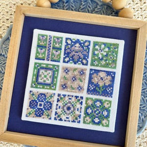May include: A framed cross-stitch sampler with nine square panels. Each panel features a different geometric pattern in shades of blue, green, and tan. The frame is light wood, and the background is a deep blue. The sampler is displayed on a blue plate.