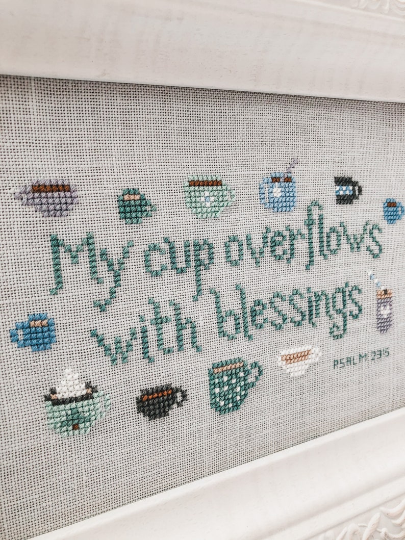 May include: Cross-stitch embroidery with a white background and teal and blue thread. The embroidery depicts a variety of coffee cups and the text "My cup overflows with blessings Psalm 23:5".