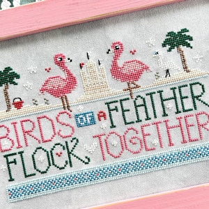 May include: Cross-stitch artwork in a pink frame. The design features two pink flamingos, palm trees, and the text "BIRDS OF A FEATHER FLOCK TOGETHER" in green and pink. A decorative piece for home decor.