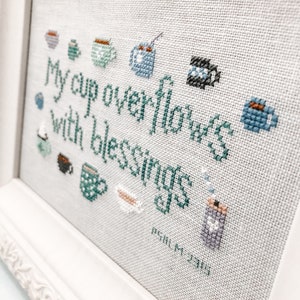 May include: Cross-stitch embroidery framed in white with a light beige background. The embroidery depicts a variety of teacups and mugs with the text "My cup overflows with blessings" and the scripture reference "Psalm 23:5".
