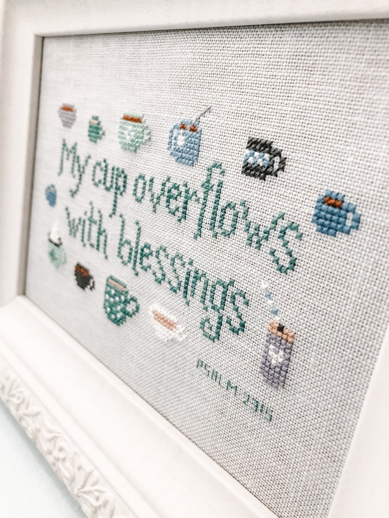 May include: Cross-stitch embroidery framed in white with a light beige background. The embroidery depicts a variety of teacups and mugs with the text "My cup overflows with blessings" and the scripture reference "Psalm 23:5".