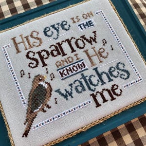 May include: Cross-stitch embroidery framed in a teal frame with a brown burlap border. The embroidery depicts a sparrow with the text "His eye is on the sparrow and I know He watches me."