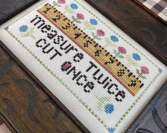 May include: A framed cross-stitch with a white background and a brown wooden frame. The cross-stitch features a yellow and brown ruler with the text "measure twice cut once" in black lettering. The ruler is surrounded by a border of green, blue, and pink flowers.