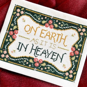 May include: A rectangular cross-stitch embroidery on off-white fabric featuring the text ON EARTH AS IT IS IN HEAVEN. The design is framed by a dark green border decorated with small pink flowers, green leaves, and gold accents.