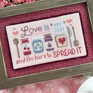 May include: Cross-stitch artwork in a wooden frame. The design features the text "Love is my jam and I'm here to spread it" with illustrations of jam jars, a spoon, a knife, and a piece of toast with a heart.