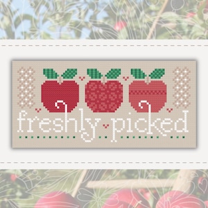 May include: A cross-stitch pattern on beige fabric featuring three red apples with green leaves arranged in a horizontal row. Below the apples, the text "freshly picked" is stitched in white, accented by small red hearts and green dots.