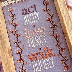 May include: Cross-stitch artwork in a wooden frame. The lavender fabric features the words "act justly, love mercy, walk humbly" in various colors. Floral vines with orange and brown flowers border the text. A white lace trim lines the inside of the frame.