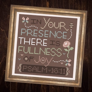 Puede incluir: Un bordado a punto de cruz enmarcado con un fondo marrón y un borde blanco. El bordado presenta un diseño floral con mariposas y abejas. El texto dice "En Tu Presencia Hay Plenitud de Alegría... Salmo 16:11".