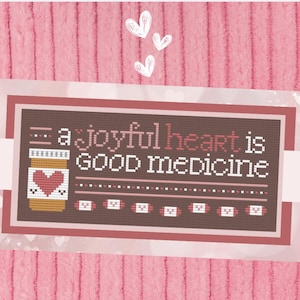 Könnte beinhalten: Kreuzstich-Design mit dem Text "a joyful heart is GOOD medicine" und einer Pillenflasche mit einem Herzen. Das Design befindet sich auf einem braunen Hintergrund mit rotem Rand, vor einem rosa strukturierten Hintergrund mit weißen Herzen.