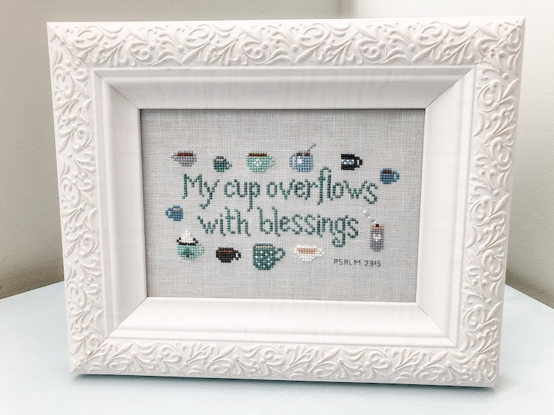 May include: White framed cross-stitch with a blue and green color scheme. The cross-stitch features a quote: "My cup overflows with blessings, Psalm 23:5".