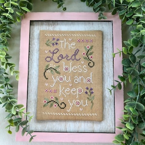 May include: A framed cross-stitch embroidery with a beige background and purple and pink floral accents. The text reads "The Lord bless you and keep you".