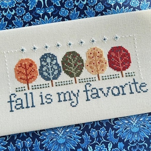 May include: A rectangular piece of light beige fabric featuring a cross-stitch design of five stylized trees in orange, blue, green, yellow, and red. Below the trees, the text reads fall is my favorite in dark blue lowercase letters.