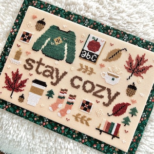 May include: Cross-stitch design with the words "stay cozy" in brown. The design includes a green sweater, a coffee cup, maple leaves, acorns, socks, and a book with an apple. The background is beige.