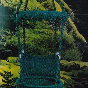 May include: A teal blue macrame hanging chair with fringe. The chair is suspended from a loop at the top and has a woven design. The chair is hanging in front of a green leafy background.