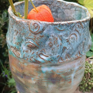Könnte beinhalten: Eine Keramikvase mit einem strukturierten, unebenen Rand und einer gesprenkelten blau-türkisen Glasur. Der untere Teil der Vase zeigt eine glatte, erdfarbene braune und beige Oberfläche mit feinen horizontalen Rillen. Orangefarbene Physalis-Stängel stecken darin.