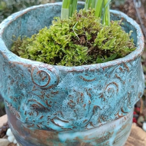 Könnte beinhalten: Ein handgefertigter Keramik-Pflanztopf mit türkisfarbener Glasur und braunen Akzenten. Der Topf ist mit grünem Moos und jungen grünen Pflanzen gefüllt. Der Pflanztopf hat eine strukturierte Oberfläche und dekorative Details.