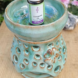 Könnte beinhalten: Ein türkisfarbener Keramik-Ölbrenner mit glänzender Oberfläche, bestehend aus einer Schale und einem perforierten Sockel mit vierblättrigem Kleeblatt-Relief. Eine kleine Glasflasche mit der Aufschrift Thymian Öl Bombafus steht in der Schale.