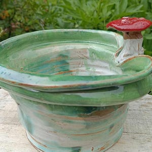 Könnte beinhalten: Eine handgefertigte Keramikschale mit leuchtend grüner Glasur und einem skurrilen roten Pilzakzent. Die Schale hat ein breites, flaches Design mit einer strukturierten Oberfläche, perfekt für dekorative Zwecke. Der Pilz hat einen roten Hut mit weißen Punkten.