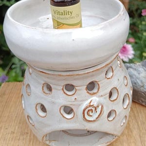 Könnte beinhalten: Ein weißer Keramik-Ölbrenner mit einer Schale oben und einem perforierten Sockel mit Spiralmuster. Eine kleine braune Glasflasche mit der Aufschrift Vitality 100% ätherisches Öl steht in der Schale. Die Keramik hat eine glänzende Glasur mit braunen Rändern.