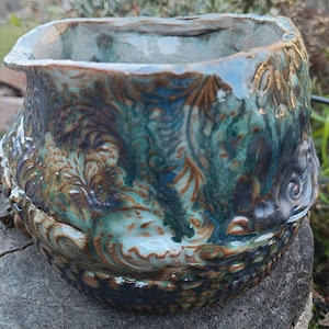 Könnte beinhalten: Ein handgefertigter Keramik-Blumentopf mit quadratischem Rand und aufwendigen, erhabenen floralen Mustern. Der Topf weist eine Mischung aus türkis, braunen und blauen Glasuren auf, die ein strukturiertes, künstlerisches Finish erzeugt. Ideal für den Innen- oder Außenbereich.