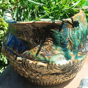 Könnte beinhalten: Ein handgefertigter Keramik-Pflanztopf mit einem strukturierten, verzierten Design. Der Topf weist eine Mischung aus Farben auf, darunter Blau, Grün und Braun, mit einem glänzenden Finish. Der Topf ist mit grünen Pflanzen gefüllt.