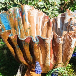 Könnte beinhalten: Eine handgefertigte Keramikschale mit gerüschtem Rand. Die Schale hat eine strukturierte Oberfläche mit einer Mischung aus brauner, blauer und weißer Glasur. Das einzigartige Design und die Farbpalette machen es zu einem dekorativen Stück für das Zuhause.