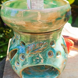 Könnte beinhalten: Ein grün und braun glasierter Keramik-Duftölbrenner mit einer breiten Schale oben und einem perforierten Sockel mit Blattmotiv. Eine kleine Flasche mit der Aufschrift Pajoma Atherisches Ol Orange steht in der oberen Schale des Brenners.
