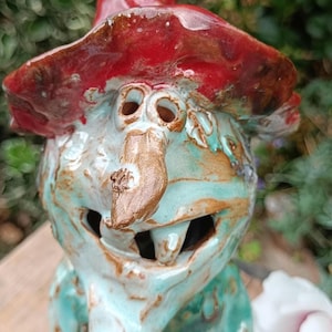 Könnte beinhalten: Eine Keramikfigur einer Hexe mit einem glänzend roten spitzen Hut und einem türkisblauen Körper. Die Figur hat eine markante braune Nase, zwei kreisförmige Augenausschnitte und einen offenen Mund, der Zähne zeigt. Die Oberfläche ist glasiert.