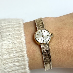 May include: A gold-toned Omega wristwatch with a round face and a mesh band. The watch has a silver dial with black hands and markers. The band is a woven design, and the watch is fastened on a wrist.