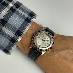 Puede incluir: Un reloj Smiths vintage con esfera plateada, bisel negro y correa de cuero negro. El reloj tiene una corona dorada y se lleva en la muñeca junto a una manga de camisa a cuadros azul y blanco.