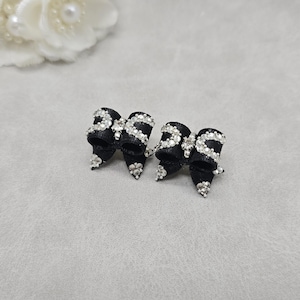 May include: A pair of black bow-shaped stud earrings featuring a textured fabric surface adorned with small, clear, sparkling rhinestones arranged in a decorative pattern across the front of each bow and along the edges.