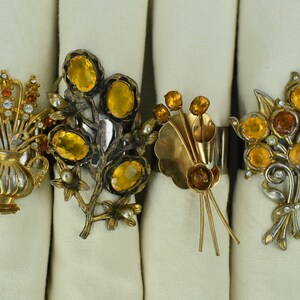 May include: Four gold and silver napkin rings with amber and clear rhinestone floral designs. The rings are on white linen napkins.