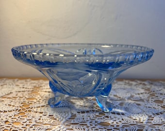 May include: A light blue, footed glass bowl with a decorative floral design. The bowl has a scalloped rim and rests on three curved feet. The glass has a slight transparency, allowing light to pass through. The bowl is likely vintage.
