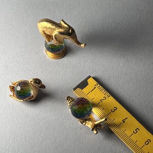 May include: Three small, gold-toned figurines: an elephant, a swan, and a snail. Each figurine features a colorful, iridescent orb. A ruler is present, indicating the snail's orb is approximately 1.5 cm in diameter.