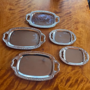 Könnte beinhalten: Fünf silberfarbene Serviertabletts in verschiedenen Größen sind auf einer Holzoberfläche angeordnet. Jedes Tablett hat eine rechteckige oder ovale Form mit dekorativen Griffen und einer reflektierenden Oberfläche. Die Tabletts scheinen aus Metall zu sein.