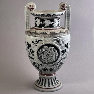 Può includere: Un vaso decorativo in ceramica con base a piedistallo, due manici e bocca larga. Il vaso presenta motivi floreali e in rilievo in bianco e nero, tra cui un medaglione centrale con un emblema dettagliato. Il vaso è alto circa 36 cm.