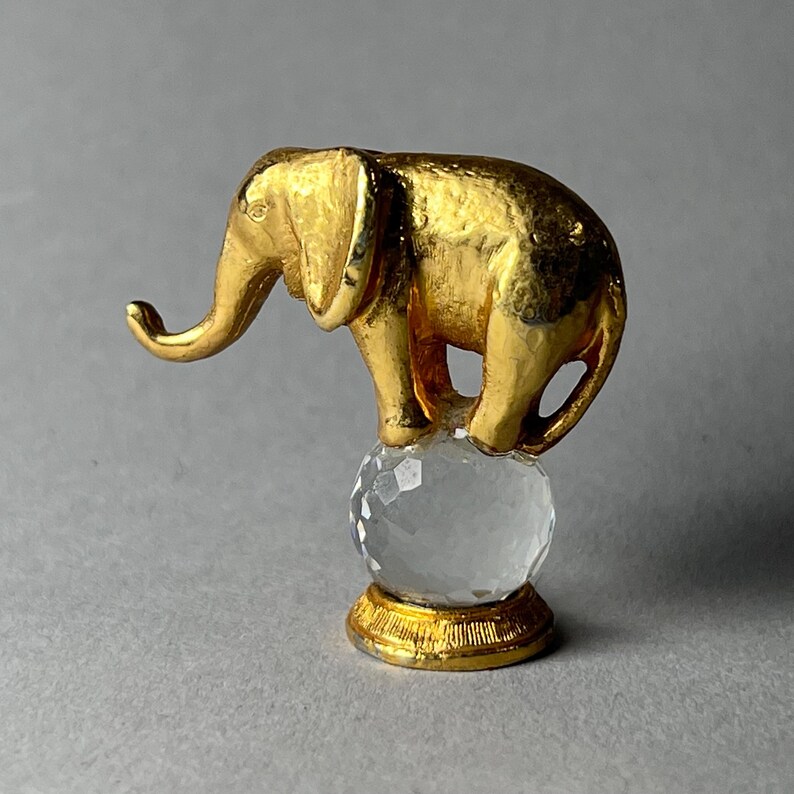 May include: A gold-colored elephant figurine standing on a clear crystal ball. The elephant has detailed features and is mounted on a gold-colored base. The figurine is set against a gray background.