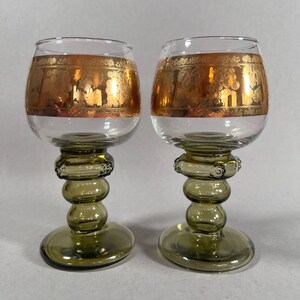 Puede incluir: Dos copas de vino vintage con cuencos transparentes y tallos verde oliva. La parte superior de los cuencos presenta una banda dorada con un diseño de uvas. Los tallos tienen un diseño bulboso decorativo y las bases son redondas.