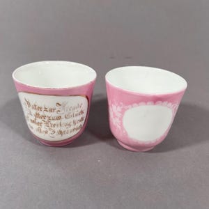 Puede incluir: Dos tazas de porcelana rosa con interiores blancos. Una taza presenta escritura dorada dentro de un borde blanco y dorado. La otra taza tiene un diseño floral blanco y un espacio ovalado blanco. Las tazas están sobre un fondo gris.