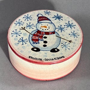 Könnte beinhalten: Eine runde Keramik-Schmuckdose mit Schneemann-Design. Der Deckel zeigt einen Schneemann mit rot-weiß gestreiftem Schal und Hut, umgeben von blauen Schneeflocken. Die Dose hat einen roten Rand und den Text "FALCONE-CALTA GIRORE".