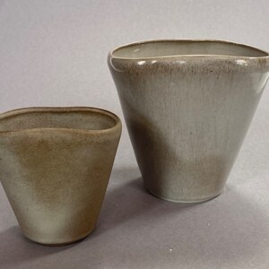 May include: Two ceramic vases in a light grey colour with a slightly textured finish. The larger vase is taller and wider than the smaller vase.