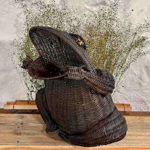 Könnte beinhalten: Ein dunkelbrauner Froschkorb aus Korbweide. Der Frosch sitzt aufrecht mit offenem Maul. Der Korb besteht aus geflochtener Korbweide und hat eine strukturierte Oberfläche. Der Frosch steht auf einer Holzoberfläche.